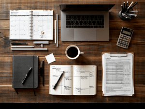 an organized wooden desk with a laptop, coffee cup, calendar, notebook, calculator, and paperwork